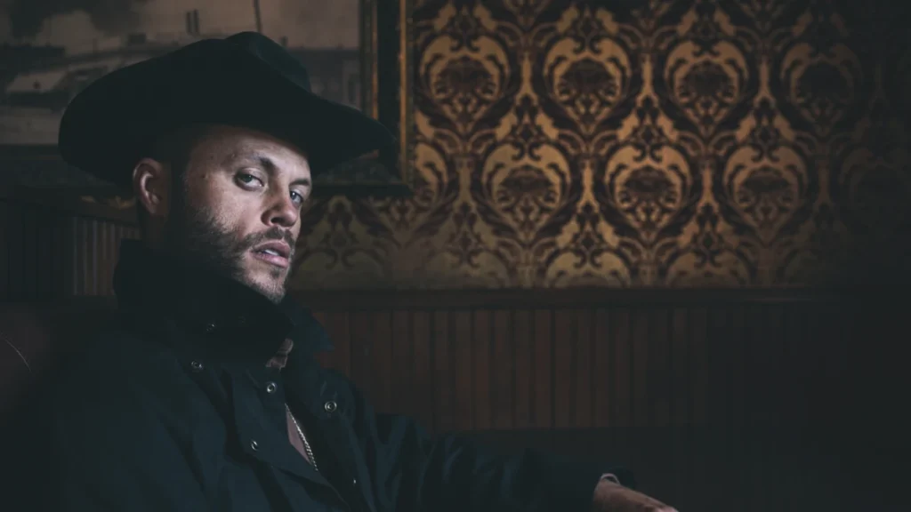 Charley Crockett in a black cowboy hat seated against patterned wallpaper