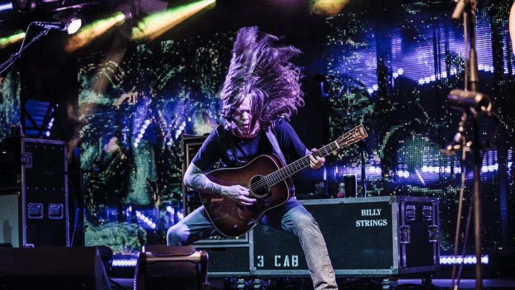 Billy Strings playing acoustic guitar on stage with his hair flying, under purple lights