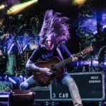 Billy Strings playing acoustic guitar on stage with his hair flying, under purple lights