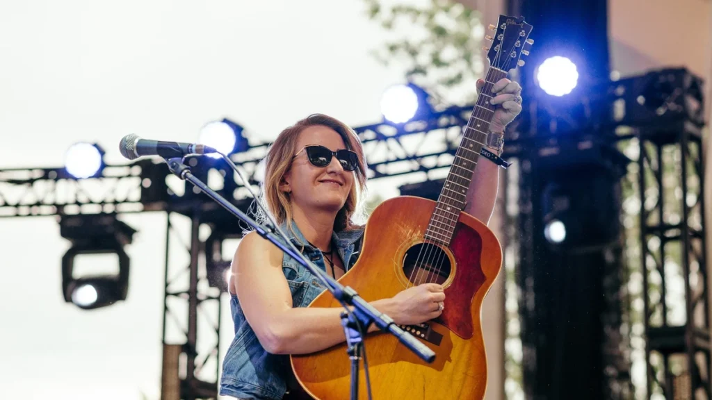 Margo Price on an outdoor stage in sunglasses, holding an acoustic guitar up by the neck